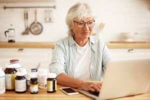 In the image, an elderly woman with white hair is using a laptop at a kitchen table. She is focused on her screen, wearing glasses and a light blue striped shirt. The kitchen has a clean, bright, and homey feel. Her bottle of Cannabiva softgels is on the table along with various other bottles of supplements, lined up beside it. She's managing her wellness plan and buying more over-the-counter CBD softgel capsules. She is also considering adding cherry-flavored CBD gummies to her order — they sound delicious.