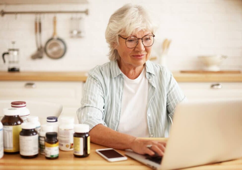 In the image, an elderly woman with white hair is using a laptop at a kitchen table. She is focused on her screen, wearing glasses and a light blue striped shirt. The kitchen has a clean, bright, and homey feel. Her bottle of Cannabiva softgels is on the table along with various other bottles of supplements, lined up beside it. She's managing her wellness plan and buying more over-the-counter CBD softgel capsules. She is also considering adding cherry-flavored CBD gummies to her order — they sound delicious.